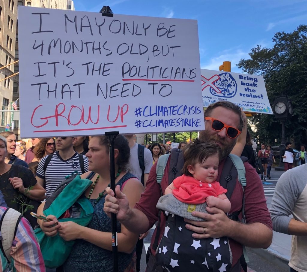 Man with baby in baby carrier holding sign with "I may only be 4 months old but it's the politicians that need to grow up"