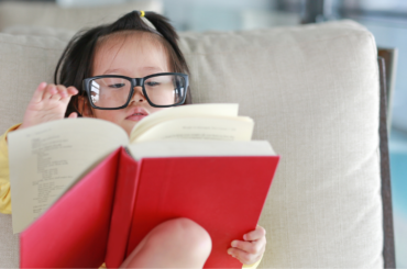 Toddler reading a big red book with a parent's glasses.