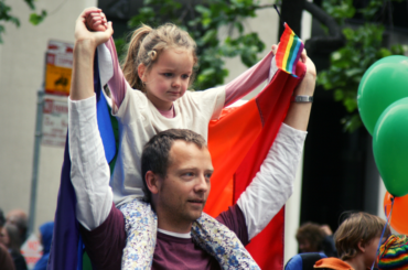 Man holding a gay pride flag carries his daughter on his shoulders.