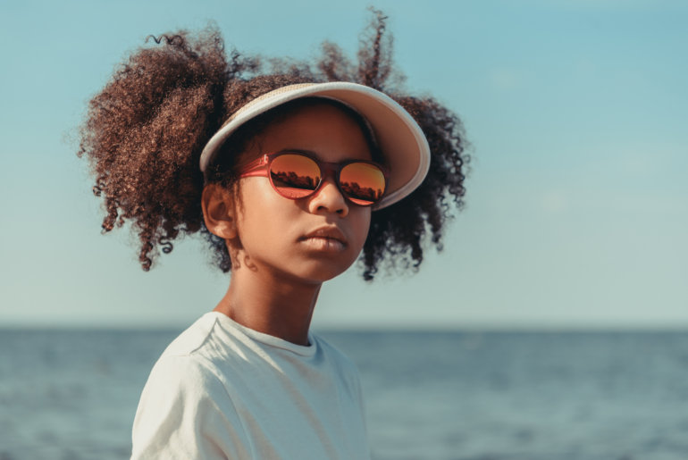 Girl on beach looking into camera through sunglasses.