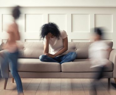 Image of mom on couch holding her head in her hand while kids run around.