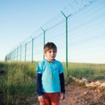 Young boy alone near tall border fence.