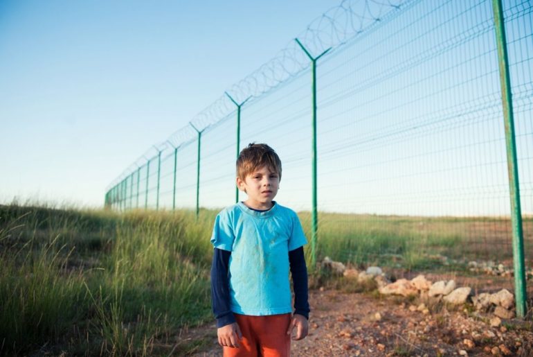 Young boy alone near tall border fence.