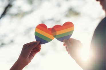 How to support an LGBTQ+ kid. Two people holding Pride hearts with sun in background.