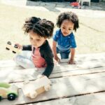 Two young kids playing with toy cars outside.