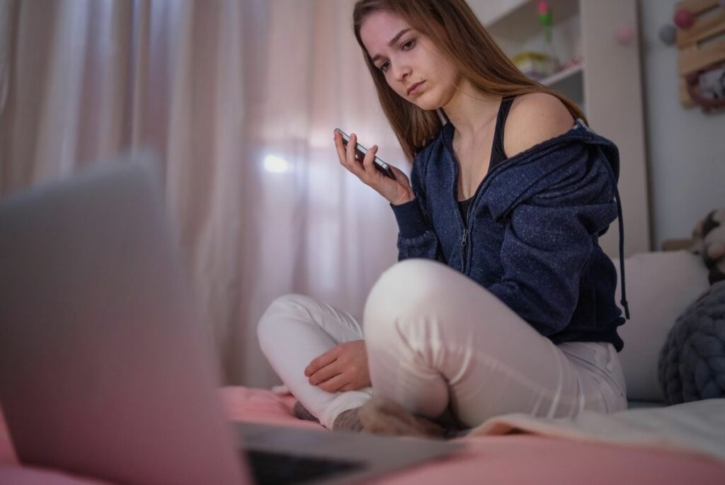 Worried teen girl sitting on bed looking at laptop.