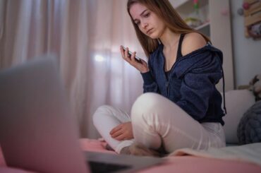 Worried teen girl sitting on bed looking at laptop.