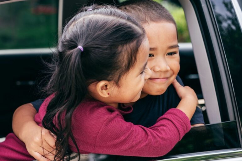 Siblings hugging and smiling in car window.