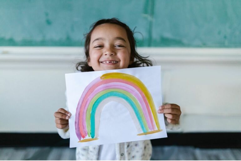 Young girl holding up rainbow painting in pre-K classroom.