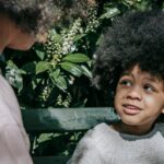 Child sitting on a bench outdoors talking to an adult.