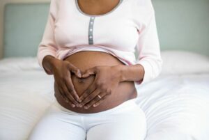 Pregnant woman sitting on bed with hands in shape of heart on her belly.