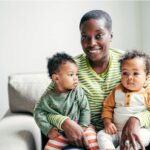 Smiling woman holding two babies on couch.