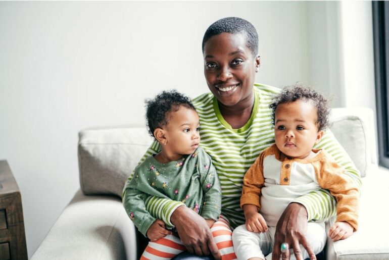 Smiling woman holding two babies on couch.