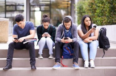 Four teens sitting outside distracted by phones.