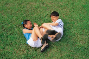 Two young Asian kids smiling while rolling around and kicking each other on a grassy lawn
