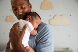 Man with dark brown skin holding a sleeping newborn baby with medium brown skin, with a white crib and cloud decorations in the background