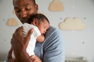 Man with dark brown skin holding a sleeping newborn baby with medium brown skin, with a white crib and cloud decorations in the background