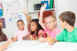 Four elementary school students sit at a table with pencils out, and two of them eagerly raise their hands