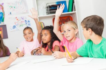 Four elementary school students sit at a table with pencils out, and two of them eagerly raise their hands