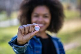 Black teen or young person wearing a denim jacket, holding out a VOTE button and smiling