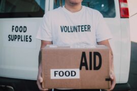Person wearing a T-shirt that says "Volunteer" holding a cardboard box that says "Food Aid"