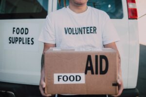 Person wearing a T-shirt that says "Volunteer" holding a cardboard box that says "Food Aid"