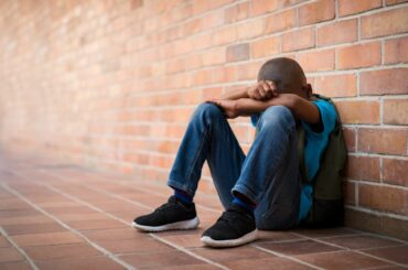 Dark brown skinned boy sitting against a brick wall, wearing a backpack, head down on his arms
