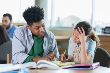 An older student tutors a younger student over a notebook and an open textbook; each holds a yellow pencil