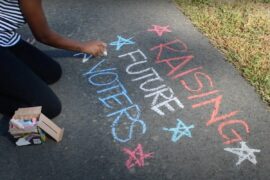 A girl writes "Raising Future Voters" on the sidewalk.