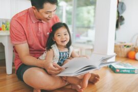 An Asian father reads a book to smiling Asian toddler on his lap