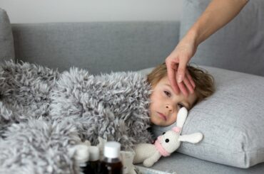 Toddler frowning under a blanket on the couch, an adult's hand checking their forehead