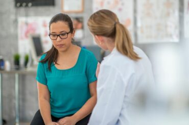 A doctor in a white coat and long blonde ponytail comforts a patient in a teal shirt, glasses, and long dark brown ponytail
