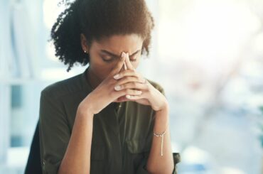 Woman with black curly hair, a green blouse, and medium brown skin has her hands folded against her forehead in a moment of concentration or stress