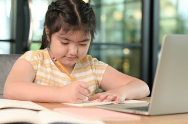 A child does homework with a pencil in front of a laptop screen