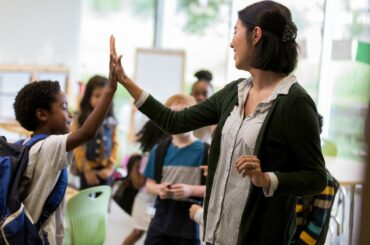 A teacher high fives an elementary school student who's wearing a backpack, with other students gathering around