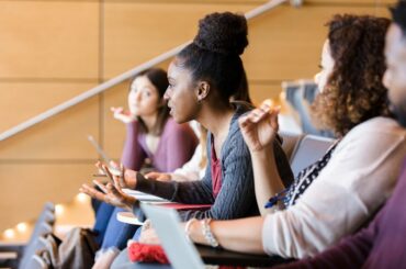 College students sit in a row of a lecture hall, and one leans forward to ask a question