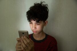 Teen with messy short brown hair, light brown skin, and striped shirt staring at a glowing phone screen in front of a white wall
