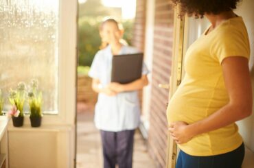 Pregnant person with yellow shirt, brown skin and curly hair holds door of home open for a midwife to enter