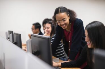 Smiling teens or young adults of many different races and ethnicities look at monitors in a computer lab