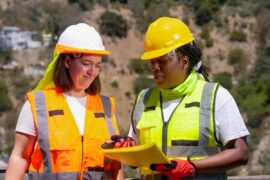 Two female workers in bright vests and hard hats look at a folder together in a dry, hot, sunny environment