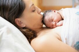 A person with medium-long brown hair lying in a hospital bed, kissing a newborn baby who is lying on their chest