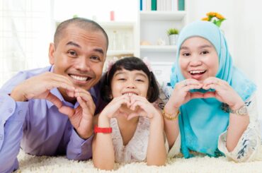 A family of three, two with dark hair and one wearing a hijab, leaning on a rug, smiling, and making hearts with their hands