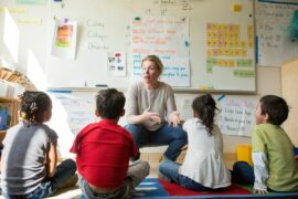 A group of first grade students sits on a rug while a teacher talks