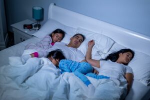 A family of four sleeps in the same bed, a toddler laying across the down comforter on top