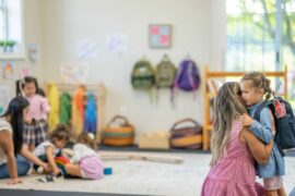 Parent hugs a small child with backpack on, as a daycare teacher plays with a few children in background