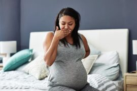 Pregnant person in gray sweats sits on a bed, her hand to her mouth in discomfort