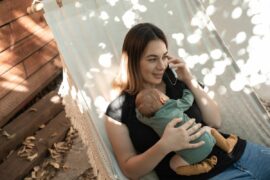 Woman with long hair holds a sleeping newborn while listening to her phone and smiling