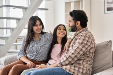 A couple sits on couch, communicating and smiling, with teen daughter sitting in between them