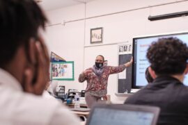 High school students look on as a teacher in a mask teaches a lesson