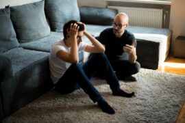A lanky teen sits against couch covering his head as a parent tries to talk to him earnestly